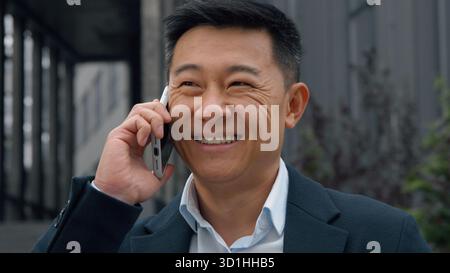 Felice sorridente uomo d'affari asiatico di mezza età che parla chiama cellulare in città uomo d'affari giapponese parla smartphone ridendo sorriso positivo Foto Stock