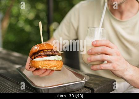 Primo piano di un uomo seduto a un tavolo da giardino con un succoso cheeseburger su un vassoio metallico che beve un latte di caffè freddo Foto Stock