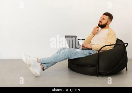 Uomo seduto su una sedia Bean Bag che utilizza un laptop mentre pensa in una camera luminosa e moderna Foto Stock