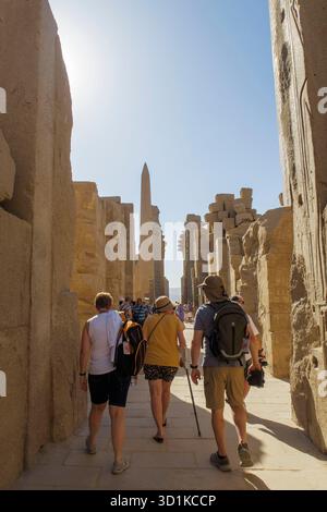 I turisti camminano attraverso un corridoio di torreggianti colonne di pietra antiche e un obelisco in un sito archeologico egizio storico Foto Stock