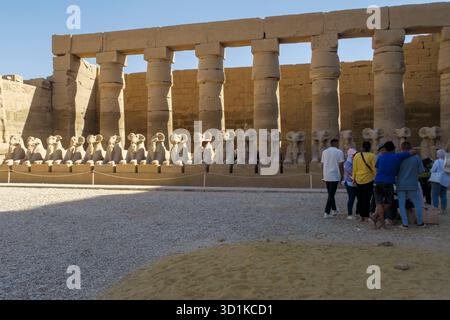Gruppo di turisti che visitano il complesso del Tempio di Karnak con una fila di statue di sfinge con testa di ariete e grandi colonne di pietra, Luxor, Egitto Foto Stock