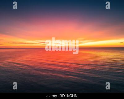 Vibrante e spettacolare tramonto sul mare o ammira l'alba sulle calme acque dell'oceano al tramonto con la superficie riflettente delle onde rosse e arancioni, vista aerea Foto Stock