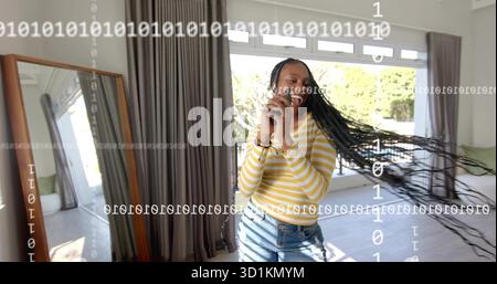 Capelli intrecciati oscillanti, donna che indossa camicia e jeans che tengono lo smartphone in salotto, con specchio Foto Stock