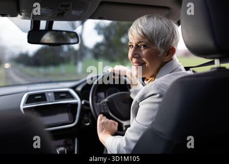 Una donna allegra ama guidare guardando indietro la fotocamera. Si trova all'interno di un'auto moderna con sfondo panoramico Foto Stock