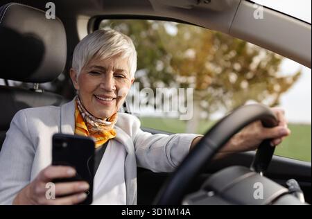 Donna anziana allegra che guarda lo smartphone in mano mentre guida un'auto, attività pericolosa e rischiosa Foto Stock