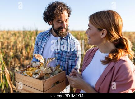 Ritratto della coppia felice che raccoglie il mais a mano. Uomo che porta una piccola cassa di pannocchie mentre guarda una bella donna accanto a lui Foto Stock