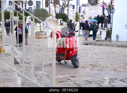 Una Vespa di colore rosso brillante si trova parcheggiata su un pendio bagnato e acciottolato, incorniciato da ringhiere di colore chiaro. Sullo sfondo, i pedoni nelle giacche e. Foto Stock