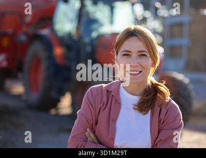 Ritratto di un bellissimo agricoltore rosso in piedi davanti al trattore sorridendo con sicurezza con le braccia incrociate Foto Stock