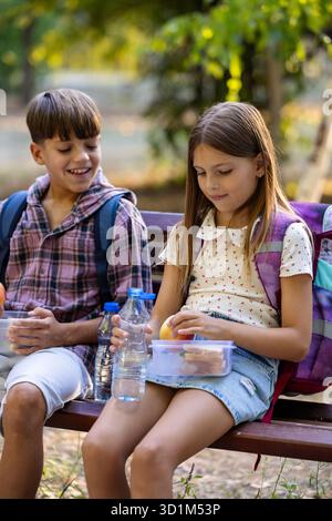 Ragazzo e ragazza della scuola media seduti su una panchina e gustati il pranzo al sacco in una giornata di sole Foto Stock