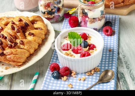 Tavolo della colazione: Ciotola di yogurt con muesli e frutta fresca: Lamponi e mirtilli, vasetti di vetro con frutta e panna montata. Foto Stock