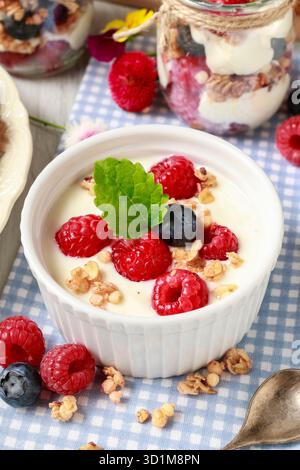 Tavolo della colazione: Ciotola di yogurt con muesli e frutta fresca: Lamponi e mirtilli, vasetti di vetro con frutta e panna montata. Foto Stock