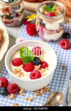Tavolo della colazione: Ciotola di yogurt con muesli e frutta fresca: Lamponi e mirtilli, vasetti di vetro con frutta e panna montata. Foto Stock