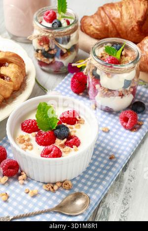 Tavolo della colazione: Ciotola di yogurt con muesli e frutta fresca: Lamponi e mirtilli, vasetti di vetro con frutta e panna montata. Foto Stock