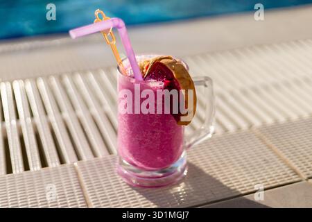 Una vivace bevanda rosa viene servita in un bicchiere trasparente, condita con una fetta di frutta tropicale e una paglia. La luce del sole splende a bordo piscina, creando un Foto Stock