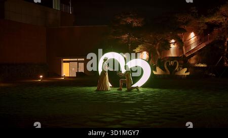 BUSAN, COREA DEL SUD - 10 OTTOBRE 2025 - uomo inginocchiato e che propone il matrimonio con una donna di fronte a una gigantesca scultura a cuore illuminato di notte Foto Stock