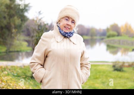 Donna anziana felice in piedi all'aperto nel parco autunnale vicino al lago con indosso caldo cappotto, cappello e sciarpa beige. Sorridente donna anziana che gode della natura durante fa Foto Stock