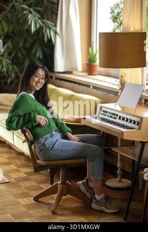 Musicista. Sorridente ragazza asiatica seduta vicino al pianoforte a casa Foto Stock