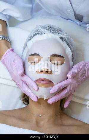Cosmetologo irriconoscibile che indossa guanti di gomma applicazione maschera foglio sul viso di donna Foto Stock