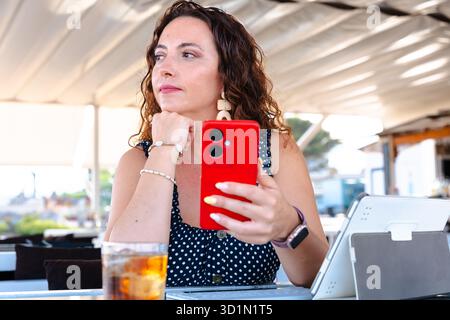 Donna con uno smartphone rosso che guarda da parte con un tablet sul tavolo Foto Stock