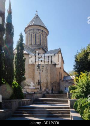 La Chiesa Kashveti di San Giorgio. Chiesa ortodossa a Tbilisi con architettura in pietra, cupola, croce, scale e vegetazione sotto un cielo azzurro. Foto Stock