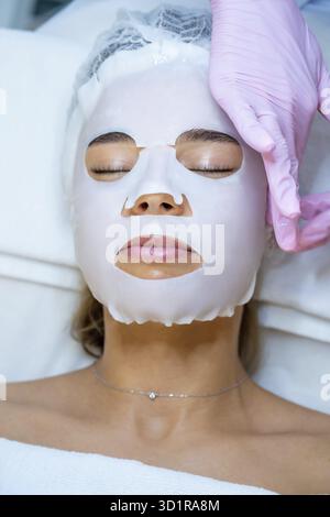 Cosmetologo irriconoscibile che indossa guanti di gomma applicazione maschera foglio sul viso di donna Foto Stock