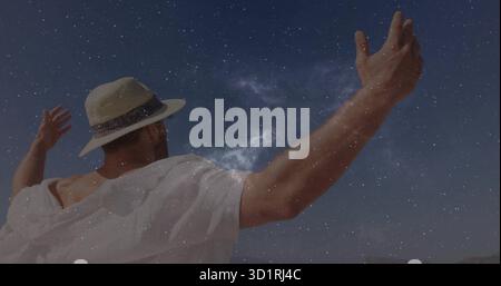 Uomo che indossa una camicia bianca sciolta che allunga le braccia sotto la via Lattea in tinta unita, con cappello di paglia Foto Stock