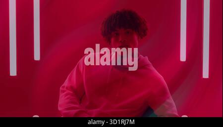 Uomo seduto con cappuccio e cuffie sovrauricolari sulla sedia in studio rosso, con tubi al neon verticali Foto Stock