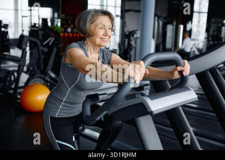 Donna anziana in sella a una cyclette in una vivace palestra Foto Stock