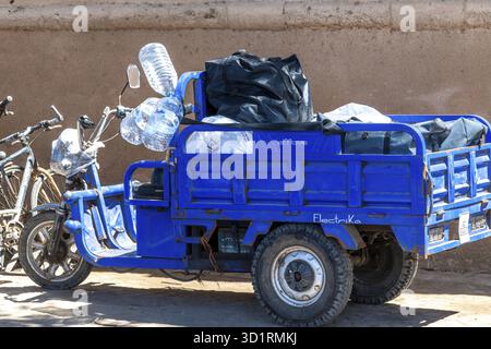 Cordova, Cordova - Spagna - 04-08-2024: Triciclo elettrico blu parcheggiato contro un muro, dotato di bottiglie d'acqua e valigia nera Foto Stock