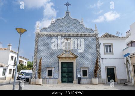 Tavira, Algarve - Portogallo - 09-18-2024: Una piccola chiesa con una facciata piastrellata a motivi blu e bianchi, porte di legno verdi e una croce sulla sommità sotto una c Foto Stock