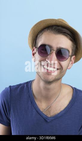 Giovane felice e sicuro di sé con cappello e occhiali da sole in piedi su sfondo blu Foto Stock