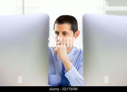 uomo penoso imprenditore d'ufficio che lavora sodo sui suoi due schermi desktop Foto Stock