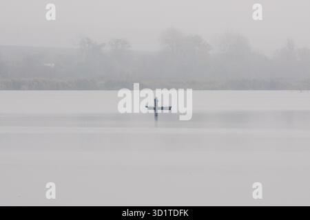 Un pescatore solitario in piedi con un'asta rotante nel mezzo di un tranquillo lago nebbioso all'alba, circondato da silenzio e da distanti sagome di alberi Foto Stock