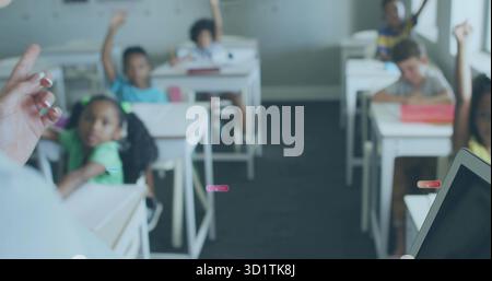 Alzare le mani gli studenti elementari seduti alle scrivanie della classe primaria di fronte all'insegnante, con un portatile Foto Stock