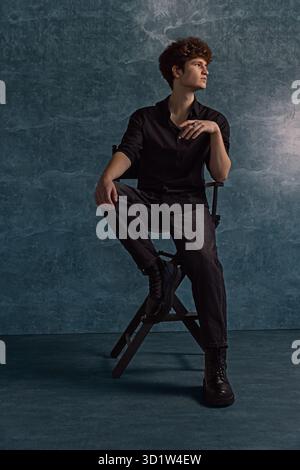 il giovane uomo siede su una sedia con una camicia nera e pantaloni scuri che guardano con attenzione verso l'alto. I suoi capelli ricci si aggiungono al suo stile unico in un ambiente da studio ben illuminato. Foto Stock