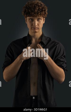 Il giovane uomo dai capelli ricci indossa una camicia nera per un look sicuro. È al chiuso su uno sfondo scuro, con particolare attenzione alla sua espressione e alla sua posa. Foto Stock