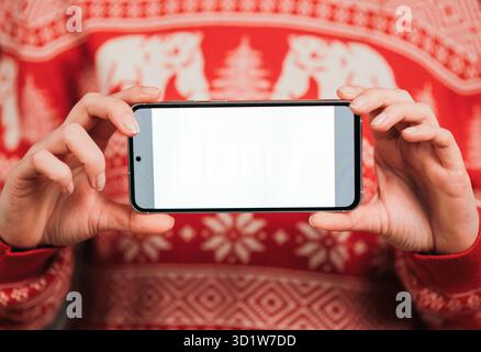 Indossa un maglione natalizio rosso che regge uno schermo orizzontale vuoto per smartphone, per i mockup e la visualizzazione delle app. Foto Stock
