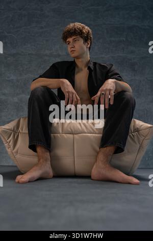 il giovane con i capelli ricci si siede comodamente su un cuscino morbido in uno studio. Indossa una camicia nera e guarda attentamente in lontananza. L'atmosfera è tranquilla e artistica. Foto Stock