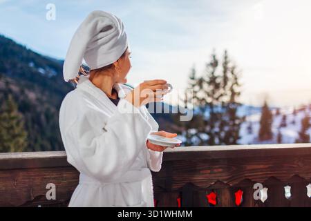 Una giovane donna beve un caffè sul balcone dell'hotel, godendosi il paesaggio invernale delle montagne, indossando un accappatoio bianco e un asciugamano sulla testa. Spazio Foto Stock
