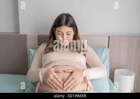 Donna asiatica incinta tiene le mani sulla pancia toccando il suo bambino curando la sua salute bella felice donna incinta tenera umore Foto Stock
