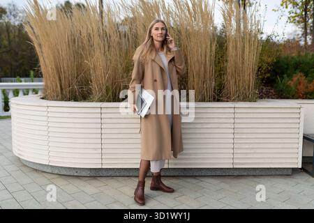 Una donna d'affari con un lungo cappotto cammina vicino a un business center, tenendo un computer portatile e parlando al telefono Foto Stock