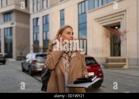 Una donna d'affari che cammina tranquillamente vicino a un moderno business center con un blocco note e un telefono in mano nelle giornate limpide Foto Stock