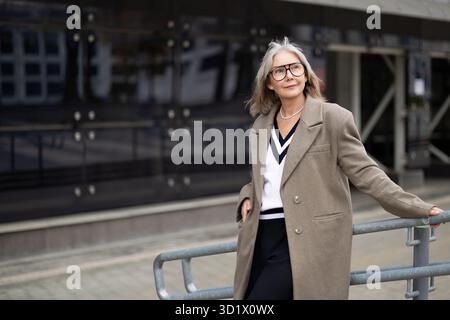 Donna anziana sicura di sé in un abbigliamento elegante si appoggia in modo informale su ringhiere all'esterno di un edificio moderno durante il giorno in una vivace città Foto Stock