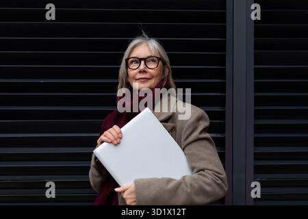 Una donna d'affari di mezza età che tiene un notebook in un ambiente urbano durante il giorno, trasuda professionalità ed eleganza Foto Stock