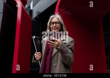Elegante donna anziana con occhiali sorride mentre tiene un telefono fuori da un moderno edificio urbano durante il giorno Foto Stock