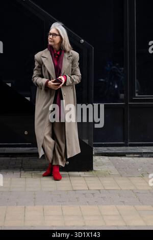 Elegante donna anziana con capelli grigi vestiti con un lungo cappotto e scarpe rosse si erge graziosamente contro una moderna parete nera in una ur Foto Stock