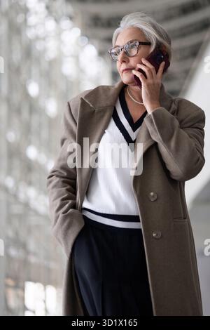 Una donna d'affari di mezza età con un cappotto elegante parla al telefono mentre si trova in un moderno edificio per uffici durante il giorno Foto Stock