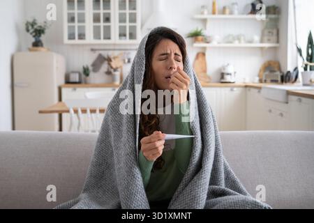 Donna malata seduta sul divano a casa con termometro nelle mani che soffre di naso che cola, starnuti Foto Stock