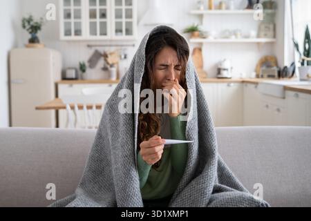 Donna malata seduta sul divano a casa con termometro nelle mani che soffre di naso che cola, starnuti Foto Stock