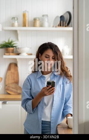 Giovane donna sorridente piacevole in piedi in cucina tenendo smartphone, guardando la ricetta video cottura Foto Stock